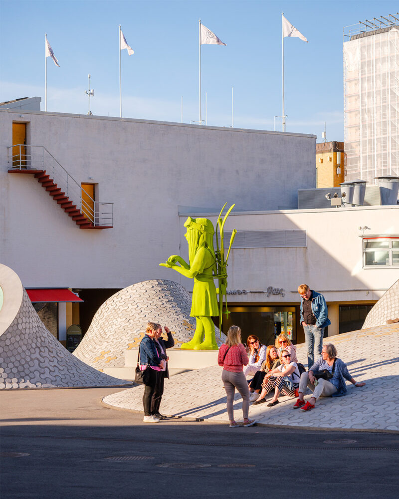 Several meters tall Wanderer giant walks on Amos Rex courtyard with skies on their back.