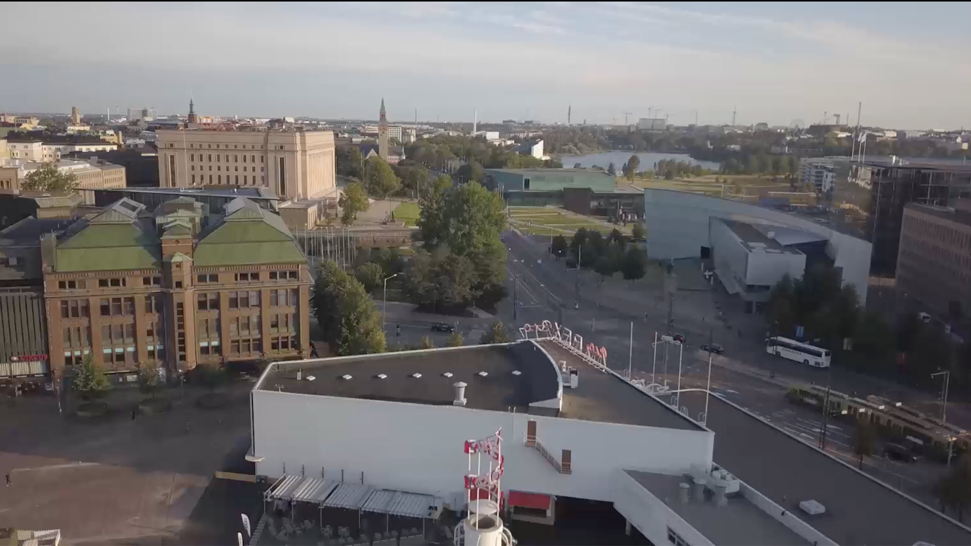 Aerial view of central Helsinki with historic and modern buildings, streets and trees, and a lake visible in the background.