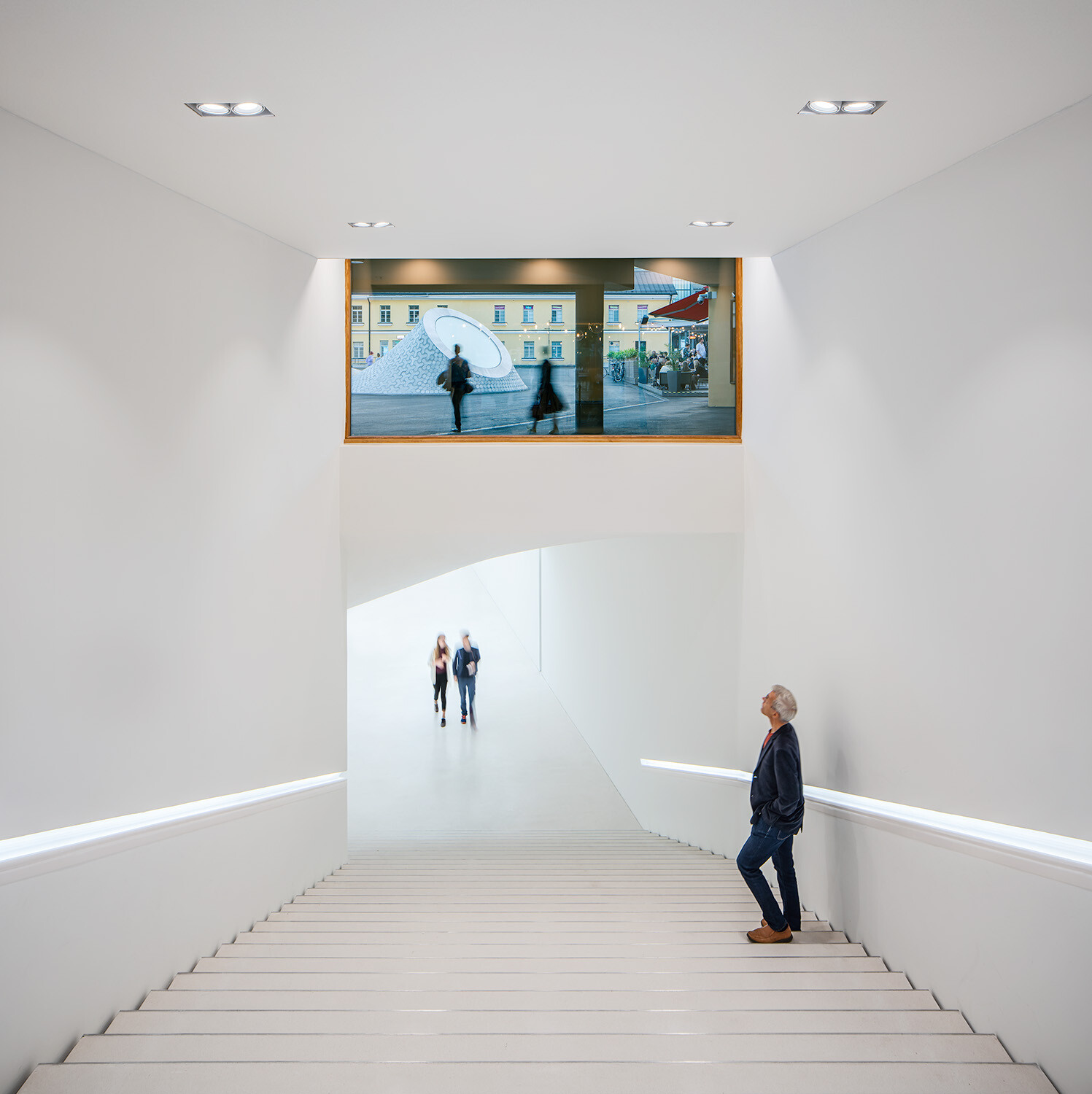 A person in black attire is standing on a wide, white staircase and looking out the window.