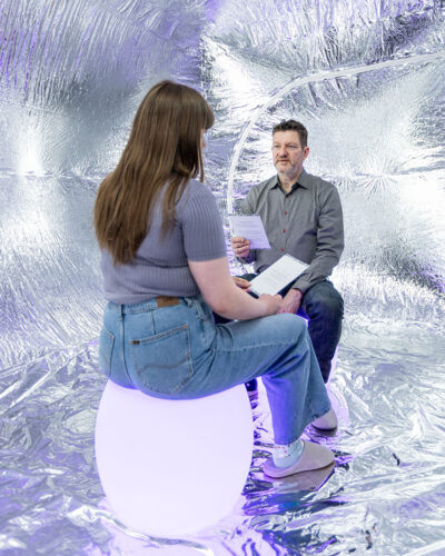 Two museum visitors sit facing each other and talk inside the Empathy Echo Chamber.