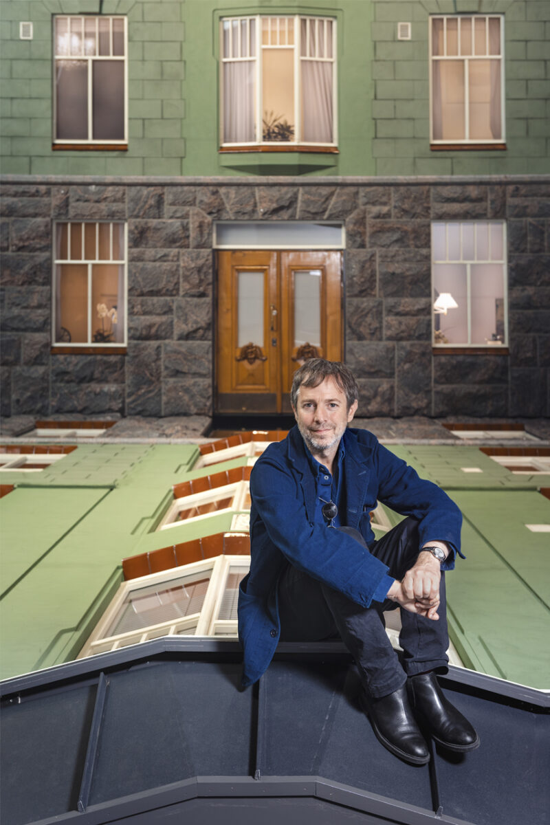The artist Leandro Erlich is sitting on his artwork Bâtiment and looking straight at the camera.