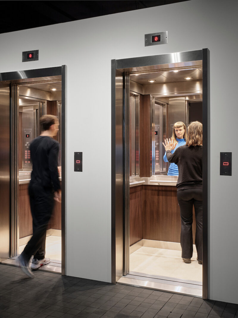 Photo showing the doorways and interiors of two elevators with people inside. The mirrors behave oddly and do not show any reflections.