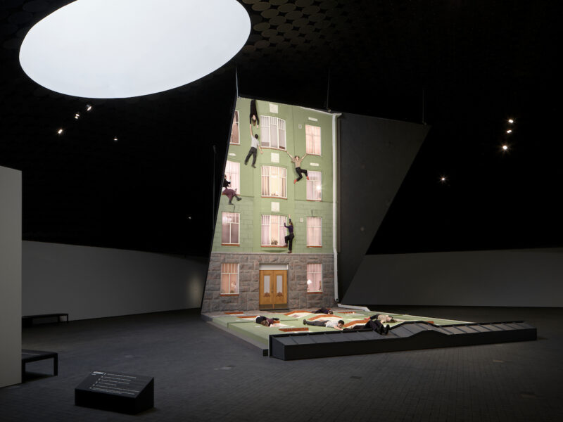 A building façade is reflected in a large mirror, creating the illusion that people are climbing on the wall.