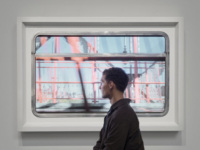 A young person stands in front of a screen showing moving views of cities and towns from different countries.