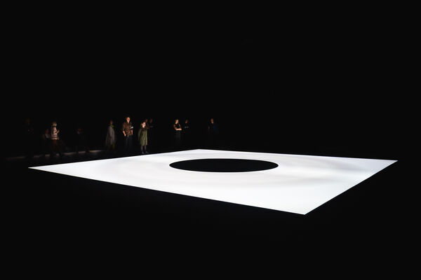 Museum visitors stand at the edge of Ryoji Ikeda’s mass. In the photo, mass appears as a gigantic luminous screen on the floor, with an expanding black hole at the center.