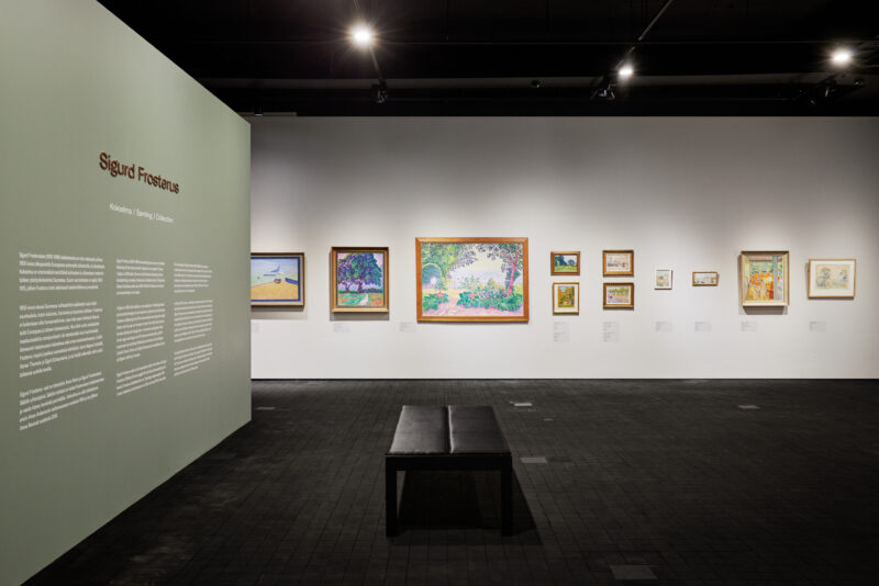 A view to the exhibition hall. There are paintings on a white wall. On the left hand side there is text on the wall. There is ablack bench in nthe middle of the room.