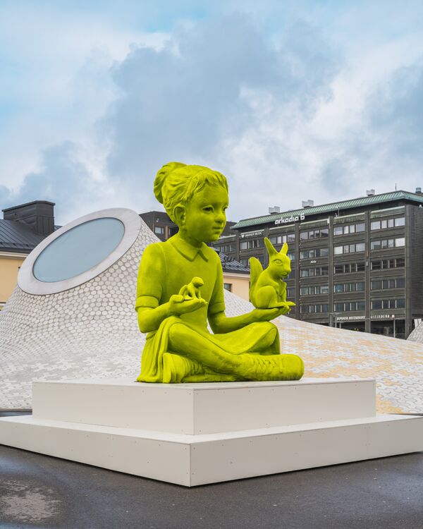 A green 3 meter tall moss girl is sitting on a pedestal on Lasipalatsi Square.