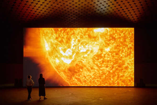 Ryoji Ikeda’s data-verse 2: the screen shows a fiery landscape resembling the surface of the Sun. A few visitors are watching and listening to the work.