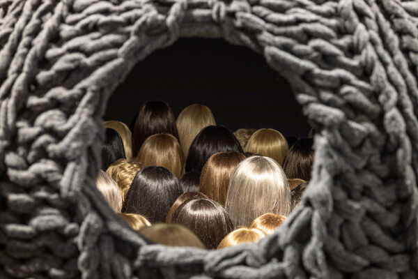 From the opening of a woven, nest-like structure resembling a giant knitted hat, countless wig-covered backs of heads peek out.