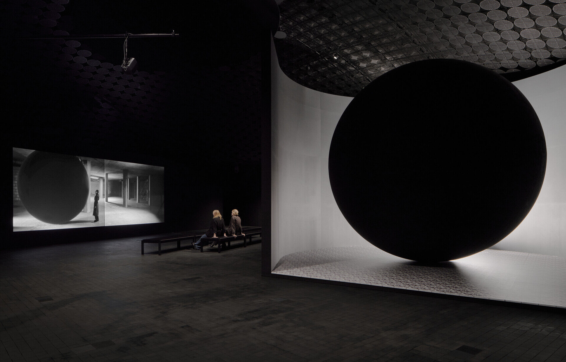 Kaksi museokävijää istuu penkillä mustan ja harmaiden värien sävyttämässä tilassa katsellen Larissa Sansourin teosta.