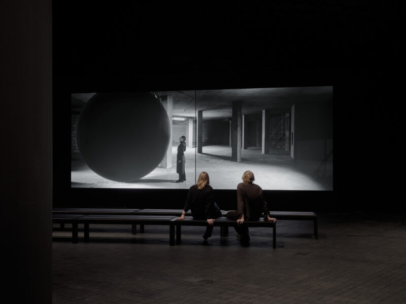 Two exhibition visitors are seated in a dark gallery, watching the video work In Vitro.