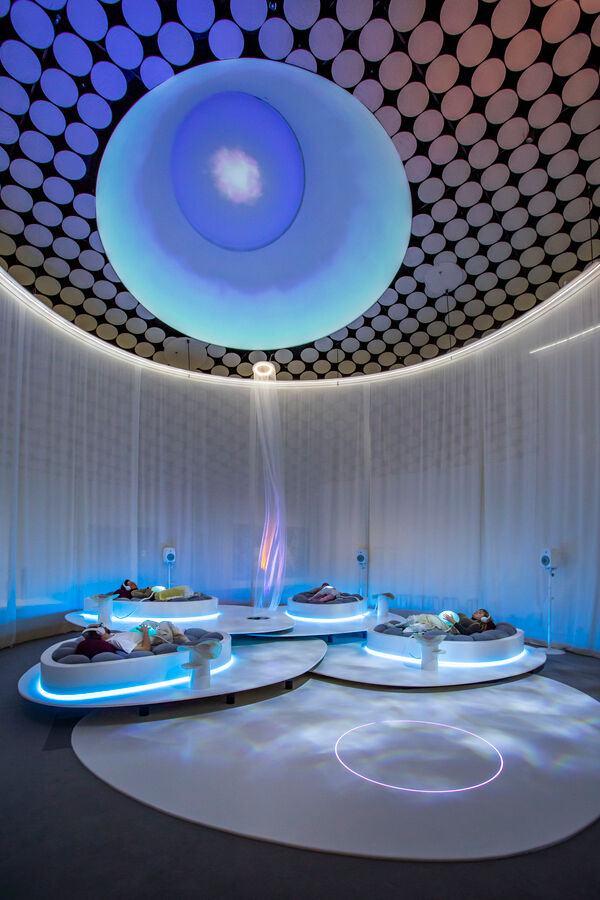 The image shows museum visitors resting on white beds inside a space surrounded by a translucent curtain, with silicone half-spheres resembling wombs placed on their stomachs.
