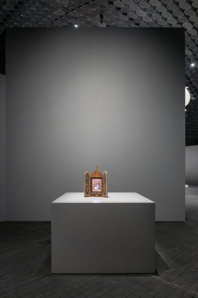 A small artwork resembling a three-dimensional icon stands on a tabletop. Inside, a small female figure is visible.
