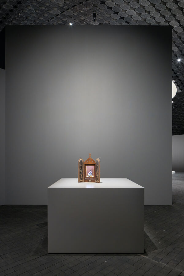 A small artwork resembling a three-dimensional icon stands on a tabletop. Inside, a small female figure is visible.