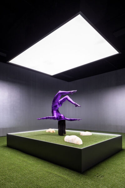 A glossy purple, gymnast-like figure appears to balance on its hair atop a pedestal, where large caterpillar-like creatures crawl across a grass-like surface.