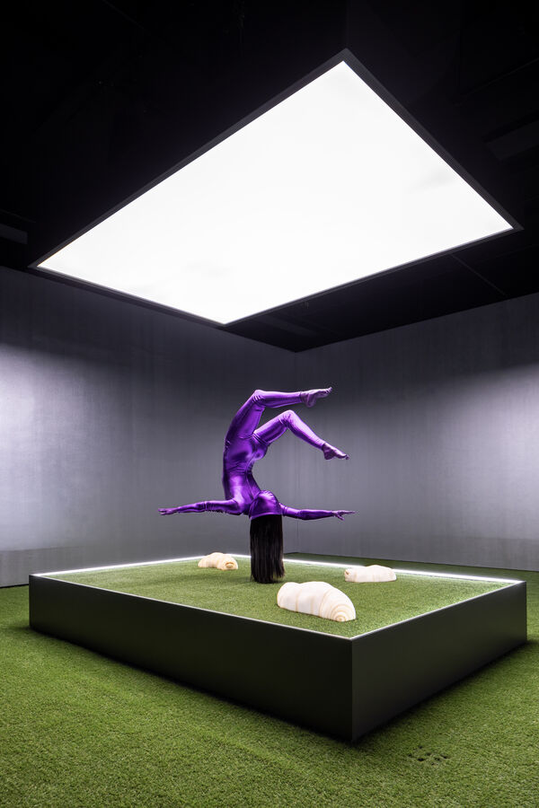 A glossy purple, gymnast-like figure appears to balance on its hair atop a pedestal, where large caterpillar-like creatures crawl across a grass-like surface.