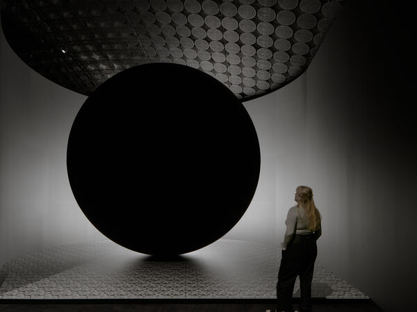 An exhibition visitor stands next to a huge sphere. The space is grey, and the sphere, larger than the visitor, resembles a black hole.