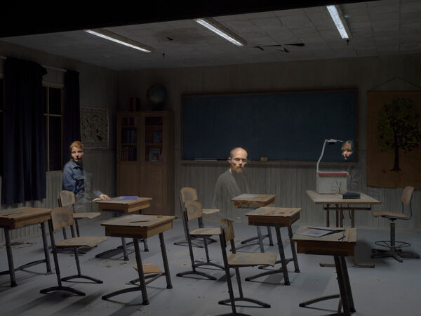 A museum visitor looks toward their reflection visible in a dimly lit, old-fashioned classroom.
