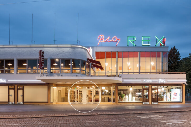 Lasipalatsens fasad i skymning, med neonskyltarna ”Bio” och ”REX” på taket. På gatunivå finns glasdörrar och stora fönster; den centrala entrén är markerad med en vit cirkel.