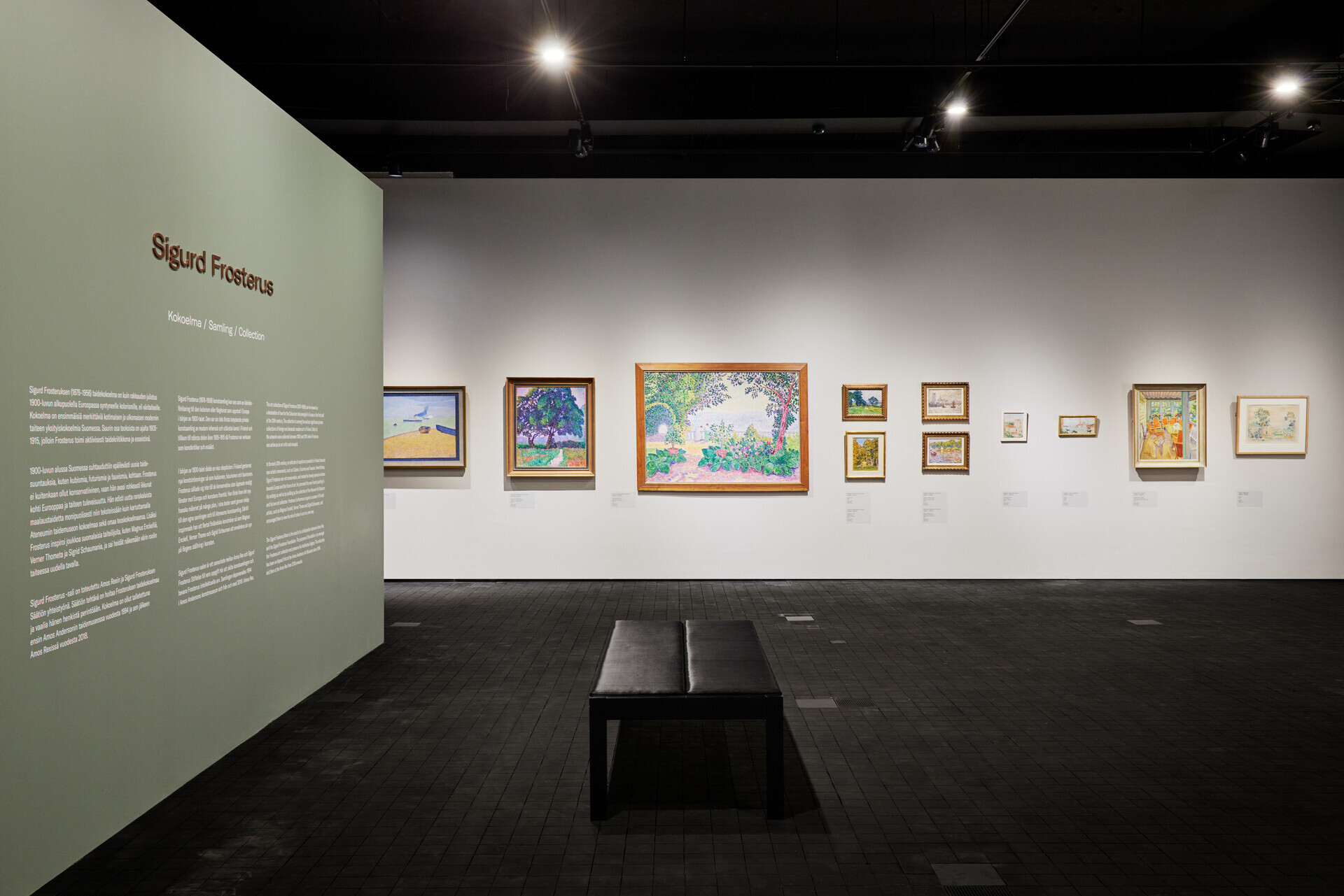 Exhibition hall with paintings displayed closely together on white walls, dark wooden block floor and a central bench for seating.