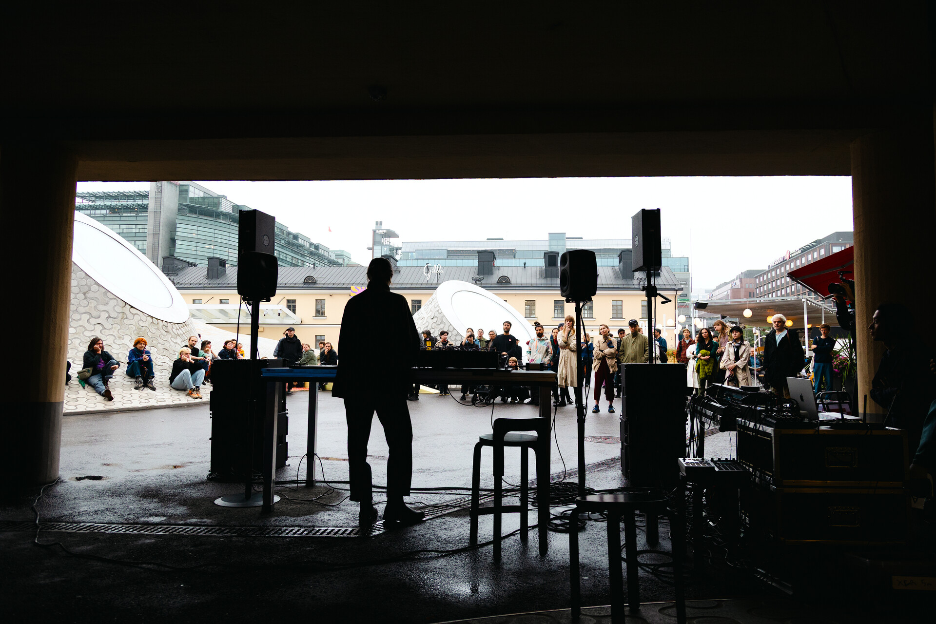 Esiintyjän siluetti äänilaitteiden äärellä katetussa tilassa, vastapäätä pientä ulkoilmayleisöä modernien rakennusten edessä harmaana päivänä.