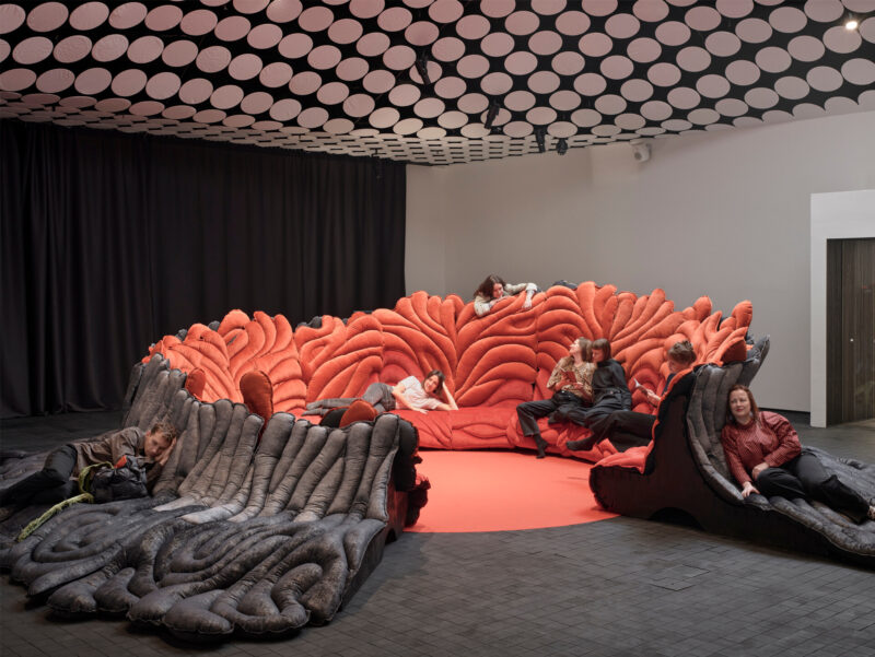 Studio Rex with a large sculptural seating structure in the centre, visitors sitting and lying on it, and a ceiling covered with a strong black-and-white graphic pattern.
