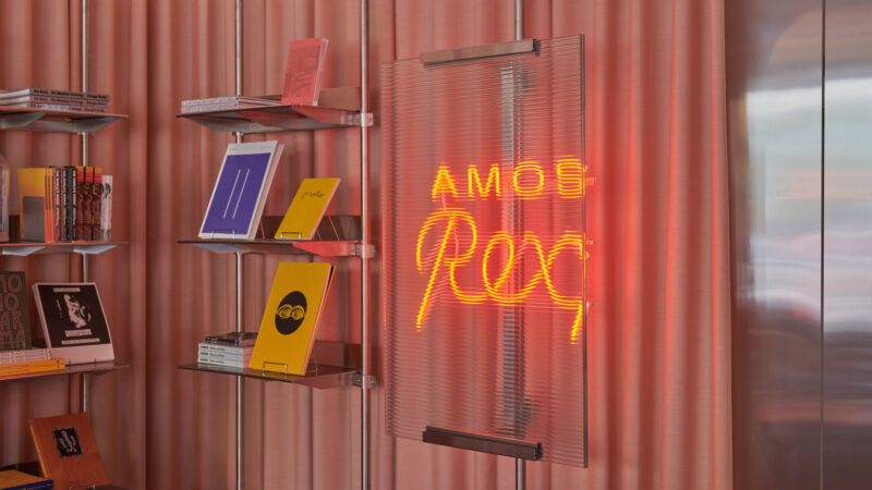 Interior of the Amos Rex museum shop with pink corrugated walls, metal shelves displaying colorful books and magazines, and a glowing orange neon sign reading “AMOS Rex.”