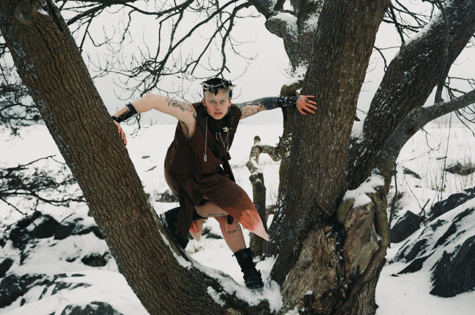 Arvi Penttilä lutar sig mot ett träd med ett vinterlandskap i bakgrunden. Konstnären bär diverse smycken och en brun tonad outfit.