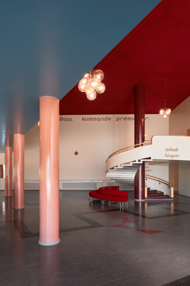 The photo shows the lobby and spiral staircase of Bio Rex. Peach-colored columns, a low blue ceiling and a higher, red ceiling.