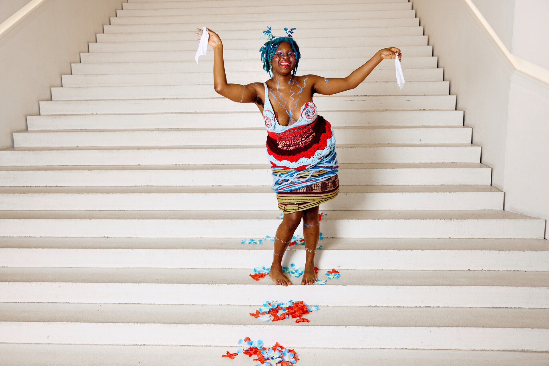 Snowdrop Áine-Fae Belmont seisoo leveillä valkoisilla portailla, pukeutuneena värikkääseen, käsintehtyyn asuun ja ilmeikkääseen meikkiin. Hän hymyilee ja pitää käsissään valkoisia kangaspaloja, ympärillään sinisiä ja punaisia koristeita portailla.