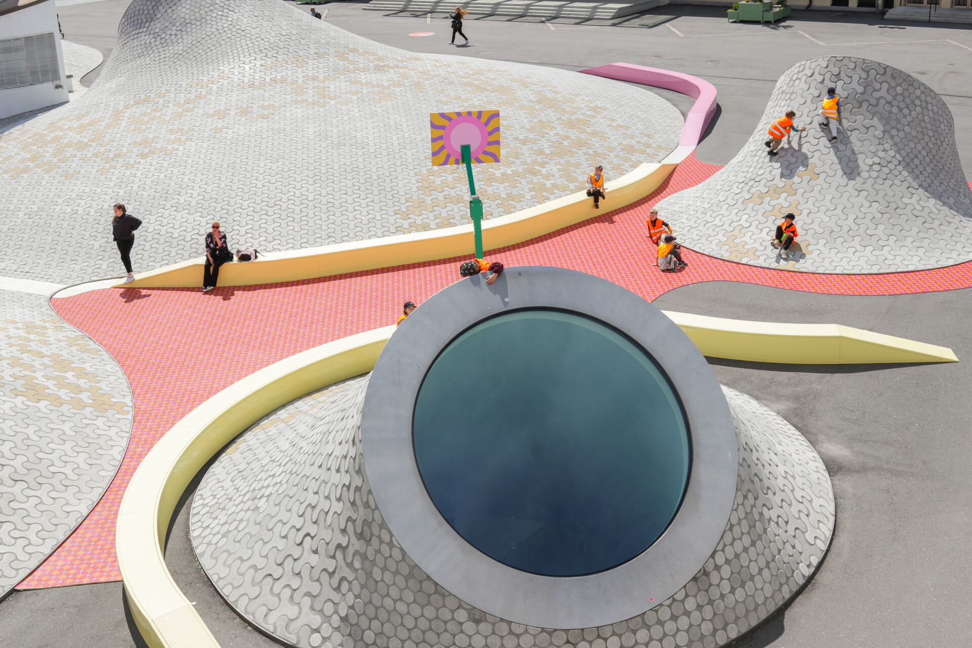 Overhead view of the Amos Rex plaza with its white, dome-shaped skylights and a circular window in the foreground. Brightly colored pathways wind between the domes, where several people, including children in orange safety vests, walk and play around a small basketball hoop.
