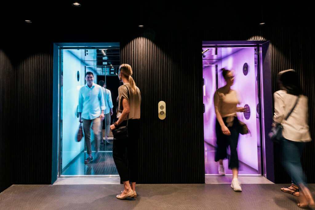 Ihmisiä astumassa ulos ja sisään kahdesta hissistä. Hisseissä on värillinen valaistus.