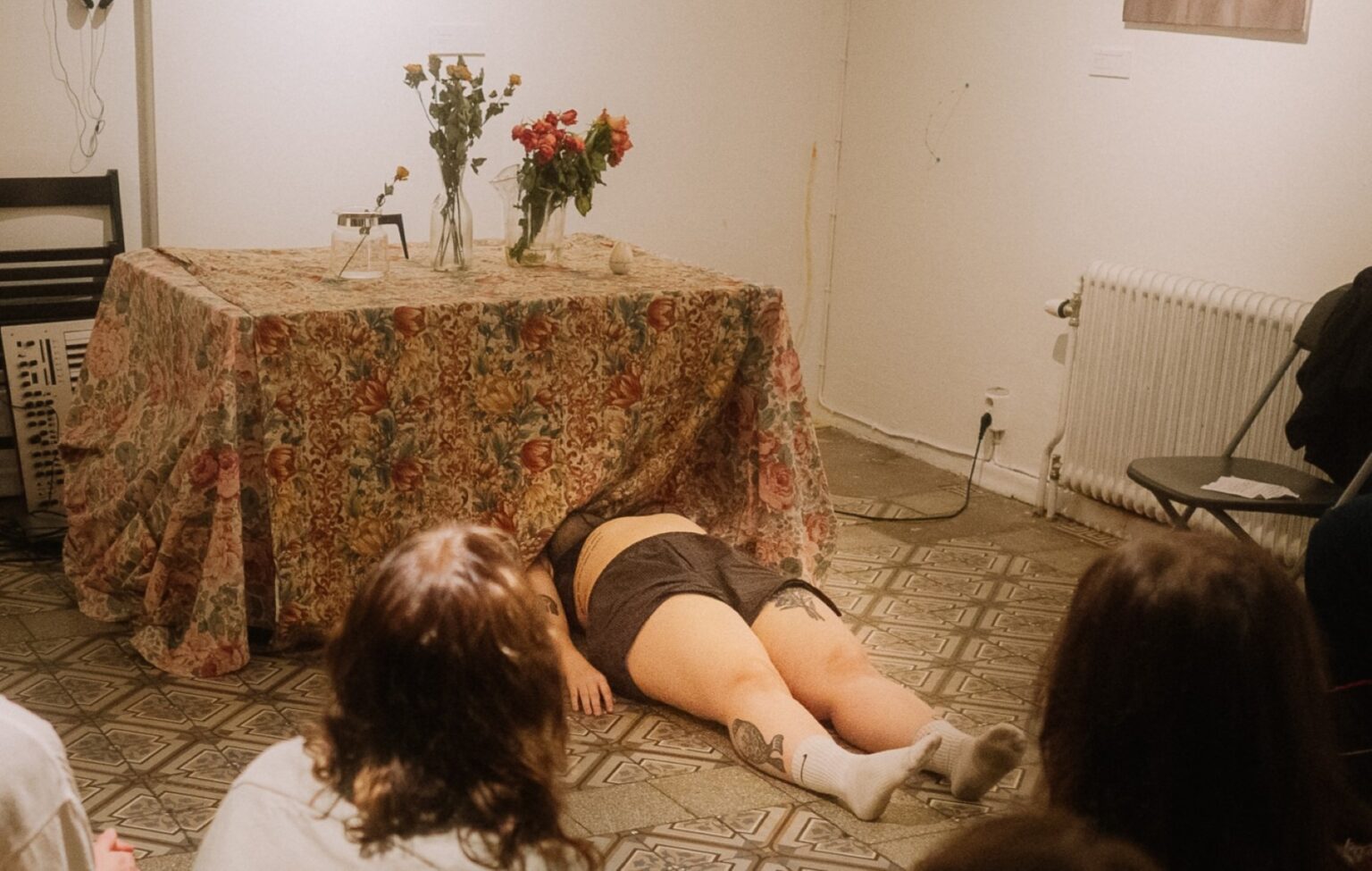 På bordet finns en blommig duk och blommor i två vaser och en kaffekanna. Under bordet ligger Vilma Tietäväinen: duken täcker hennes överkropp.