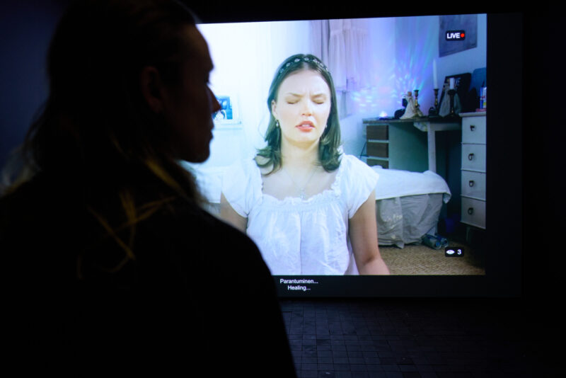 Screenillä näkyy video valkopaitaisesta naisesta, jolla on silmät kiinni. Etualalla näkyy näyttöä katsovan henkilön siluetti.