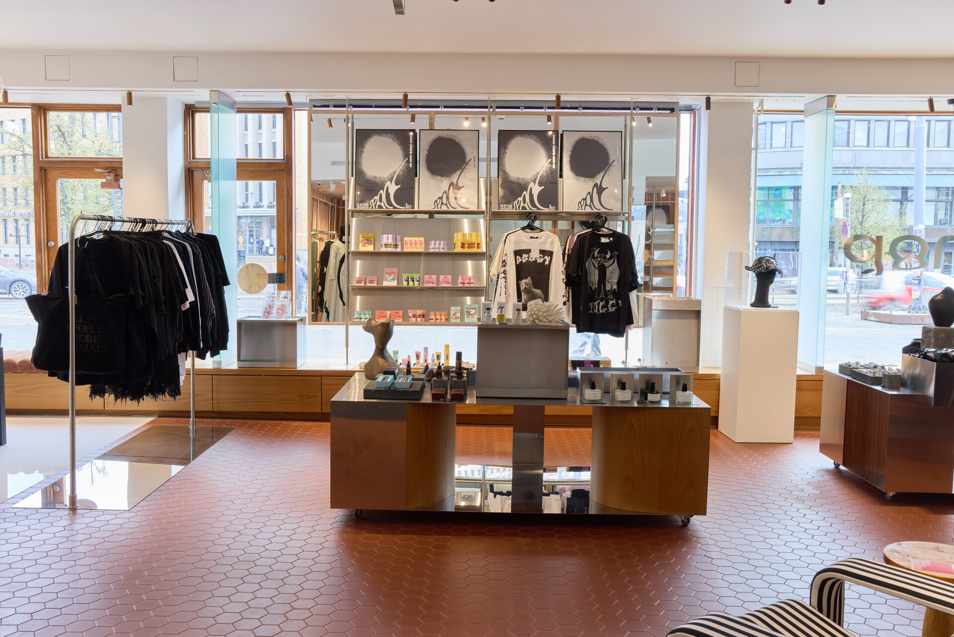 Interior of the Amos Rex shop featuring the SS26 collection. A central display table holds beauty products and small objects, flanked by racks of black clothing and shelves with colourful packaged goods. Graphic black-and-white printed T-shirts hang on the back wall alongside large art prints. The space has terracotta hexagonal floor tiles, warm wood fixtures, and large windows overlooking the street.