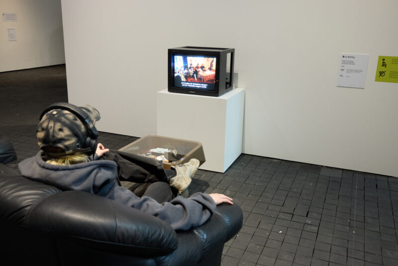 Mustalla nahkasohvalla istuu hahmo kuulokkeet päässä. Hän katsoo vanhanaikaista televisiota, jossa pyörii video. Television ja sohvan välissä on sohvapöytä.