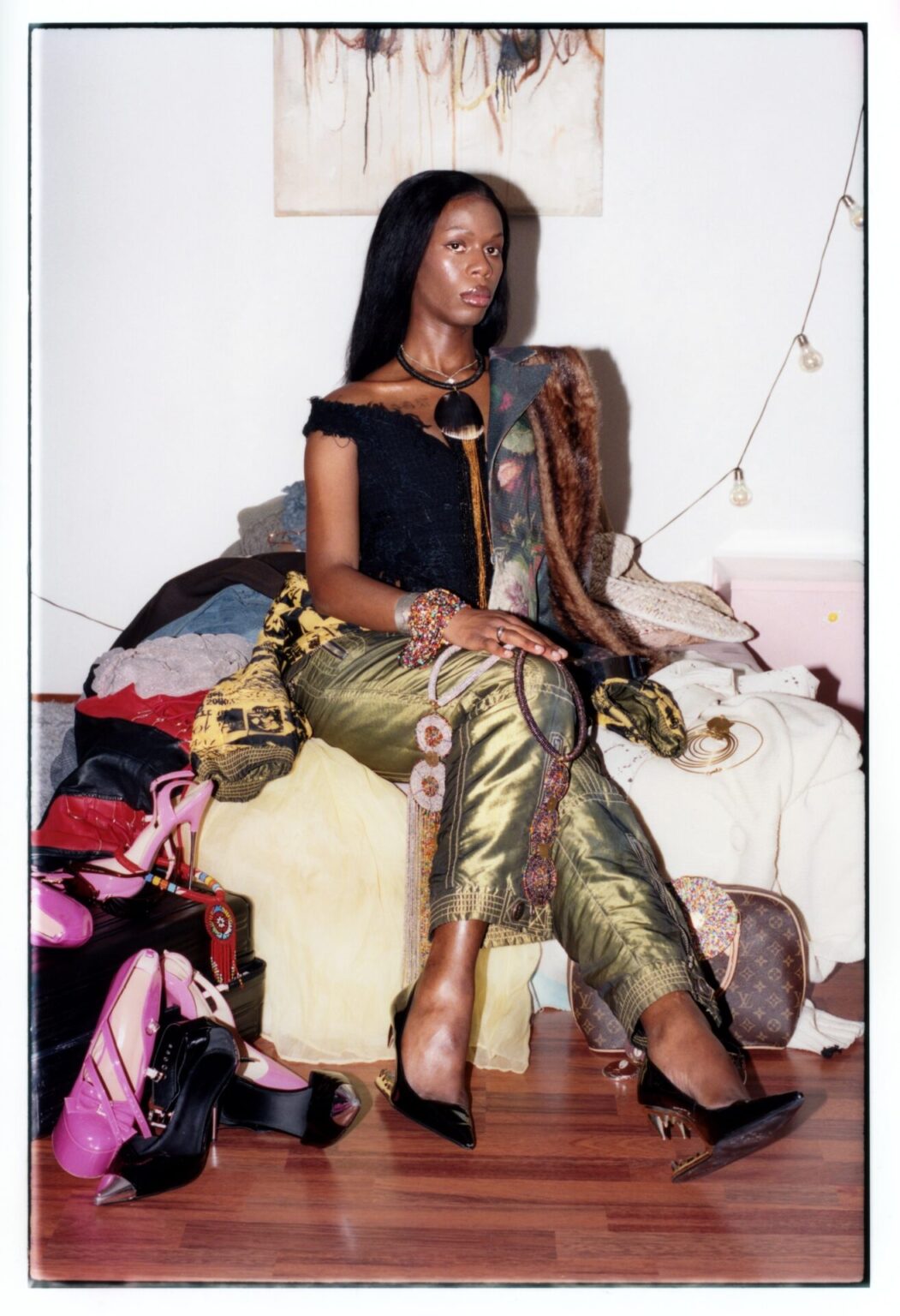 Porträtt av Angela Mugisha, konstnären bakom Money Cowry, klädd i en svart topp och olivgröna byxor, sittande på en säng omgiven av en mängd väskor och accessoarer.