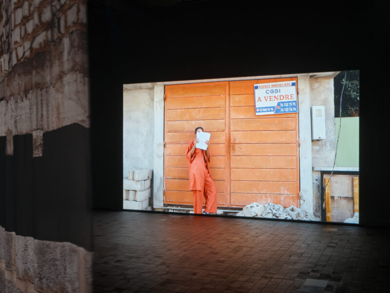 ett mörkt utställningsrum syns en stor bild av en orangebrun byggnad med en orange klädd människofigur framför.