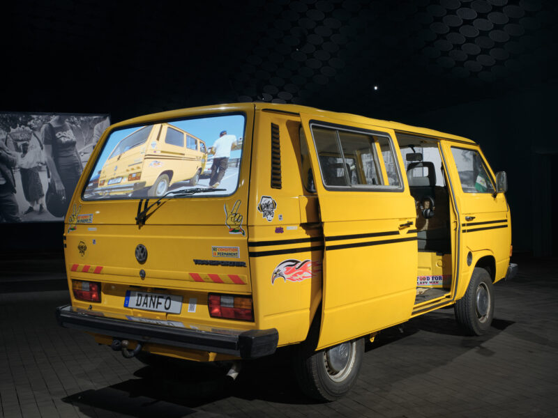A bright yellow van stands in a dimly lit exhibition gallery with its side door open.