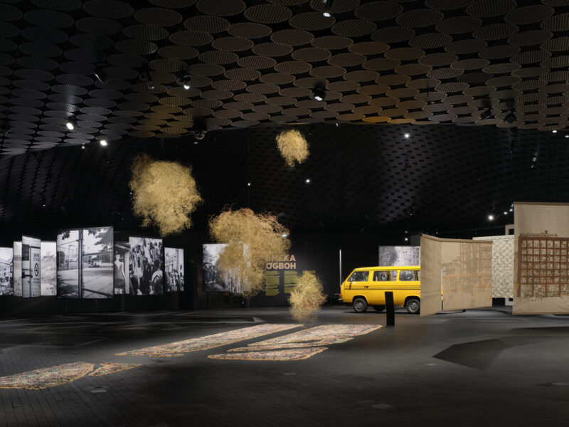 A dark exhibition hall features colourful floor artworks, suspended golden sculptures and a yellow van in the background.